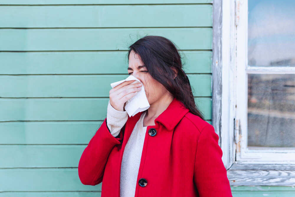 Młoda kobieta w czerwonym płaszczu trzyma chusteczkę koło nosa i kicha